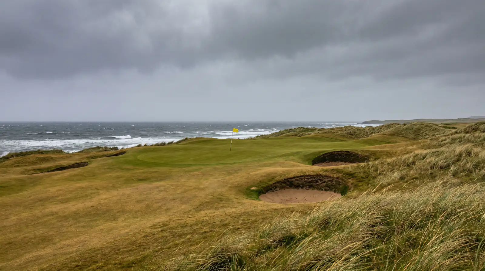 Coastal links golf course with wind-blown pin flag on a championship par four near the sea